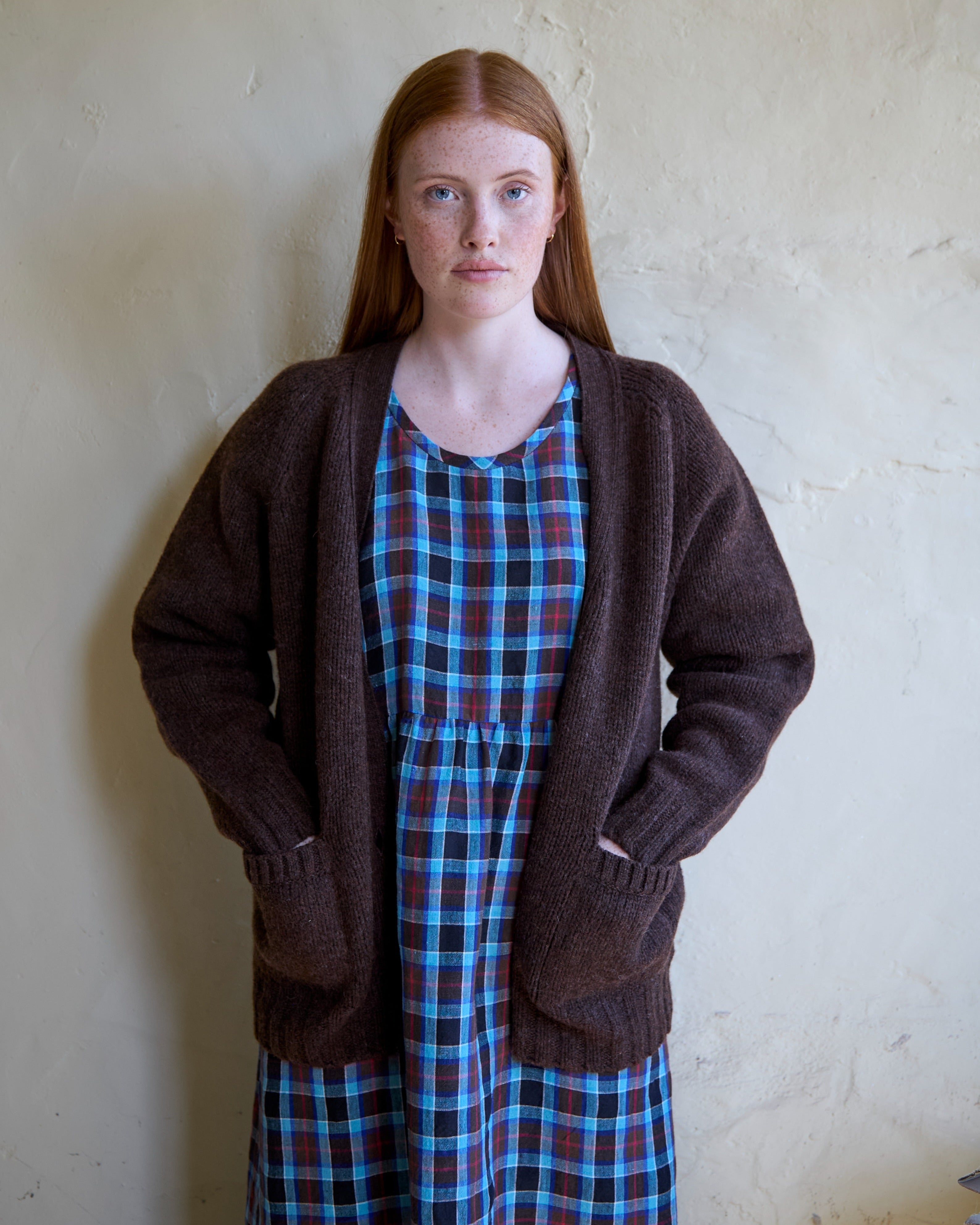 Person wearing a brown cardigan over a plaid dress against a plain wall